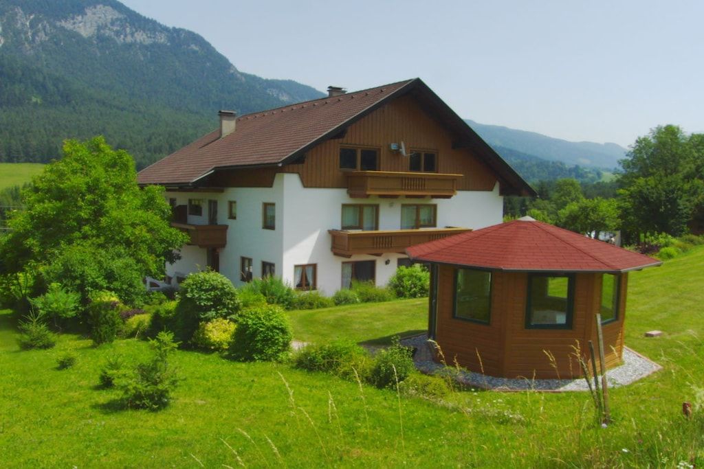 Une maison à deux étages, avec un toit rouge, entourée d’une végétation luxuriante et avec des montagnes en arrière-plan.