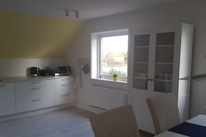 A kitchen with white cabinets, a microwave, and a coffee maker. There's a window with a view of a house and a plant on the windowsill. - Holiday apartment with 1 bedroom (Hohwacht)
