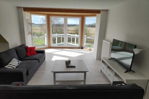 A living room with a sofa, a coffee table, a TV on a stand, and a view through large glass doors to the outdoors. - Holiday apartment with 1 bedroom (Hohwacht)