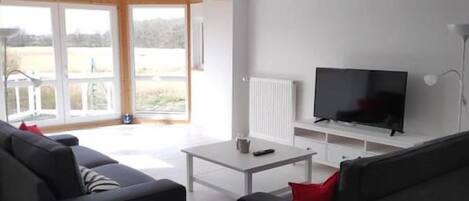 A modern living room with a flat-screen TV, two sofas, a coffee table, and a sliding glass door leading to a balcony.