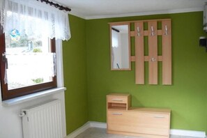Een kamer met een radiator, een raam met een witte gordijn, een houten garderobe met spiegel, en een klein houten kastje.