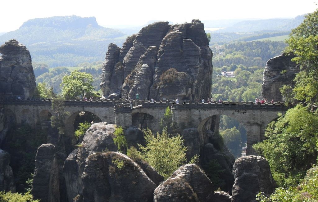 Un puente de piedra con múltiples arcos que cruza un profundo desfiladero rocoso, rodeado de abundante vegetación y colinas al fondo.