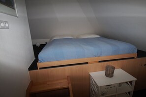 Ein Einzelbett mit Holzrahmen und einer blauen Bettdecke in einem Raum mit weißen Wänden und einem kleinen weißen Tisch mit einem Korb darauf.