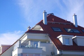 Ein Wohngebäude mit rotem Ziegeldach, mehreren Schornsteinen und einem Balkon mit Geländer.