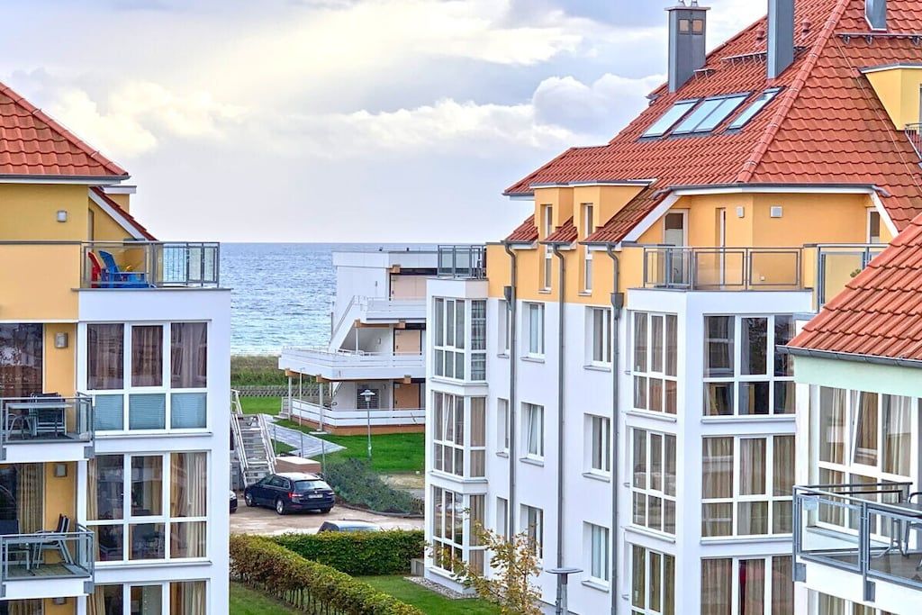 A multi-story residential building with balconies, a car parked in front, and a view of the sea.