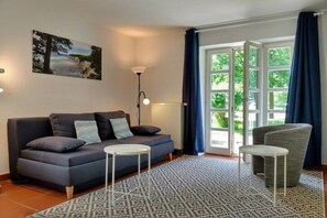 A modern living room with a sofa, two side tables, a patterned rug, and a large window with curtains. - Holiday apartment with 1 bedroom (Dranske)
