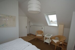 Ein Schlafzimmer mit einem Bett, zwei Korbstühlen, einem kleinen Tisch, einer Karte an der Wand, einem Kreuz an der Tür und einem Fenster.