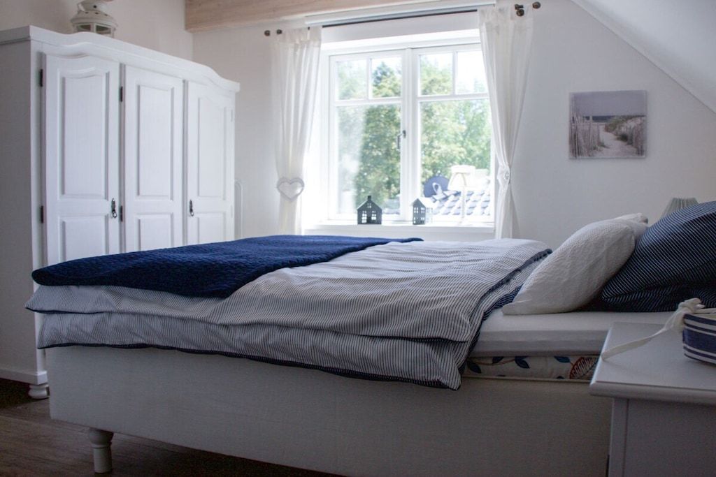 A bedroom with a bed, a window with curtains, a white wardrobe, and a small table with a lamp.