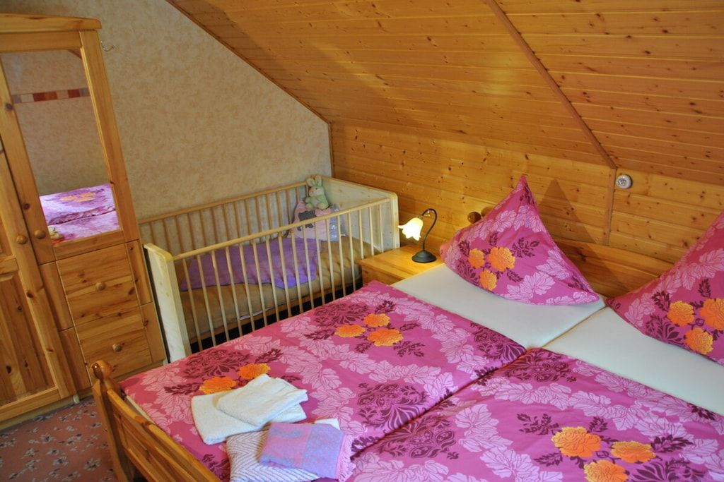 Een kamer met een houten plafond, een ledikant met een baby, een bed met bloemenbeddels en een nachtkastje met een lamp.