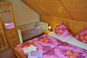 Une pièce avec un plafond en bois, un berceau où se trouve un bébé, un lit avec une literie à motifs floraux et une table de chevet avec une lampe.
