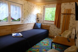 Quarto com cama, uma pequena mesa, uma cadeira, janela com cortinas e parede com padrão de folhas.