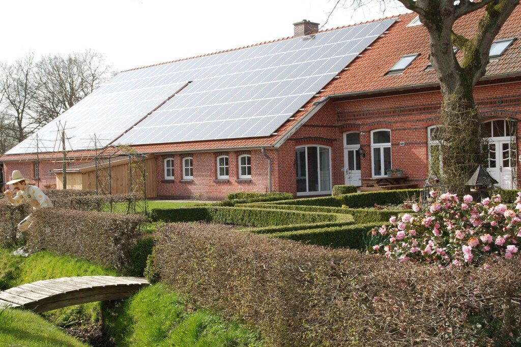 Une maison en briques rouges, avec un toit en pente, des panneaux solaires, un jardin bordé de haies et un pont en bois.