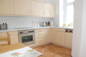 Een keuken met lichte houten kastjes, een kookplaat, oven en een eettafel met een bloemrijk tafelkleed.