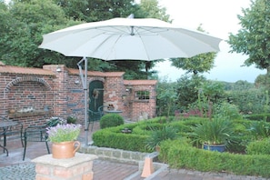 Een terras met een bakstenen muur, een witte parasol en een bakoven.