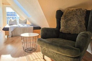 Ein gemütliches Zimmer mit einem grünen, gepolsterten Sessel, einem runden Beistelltisch aus Draht und einem Bett mit weißem Kopfteil.
