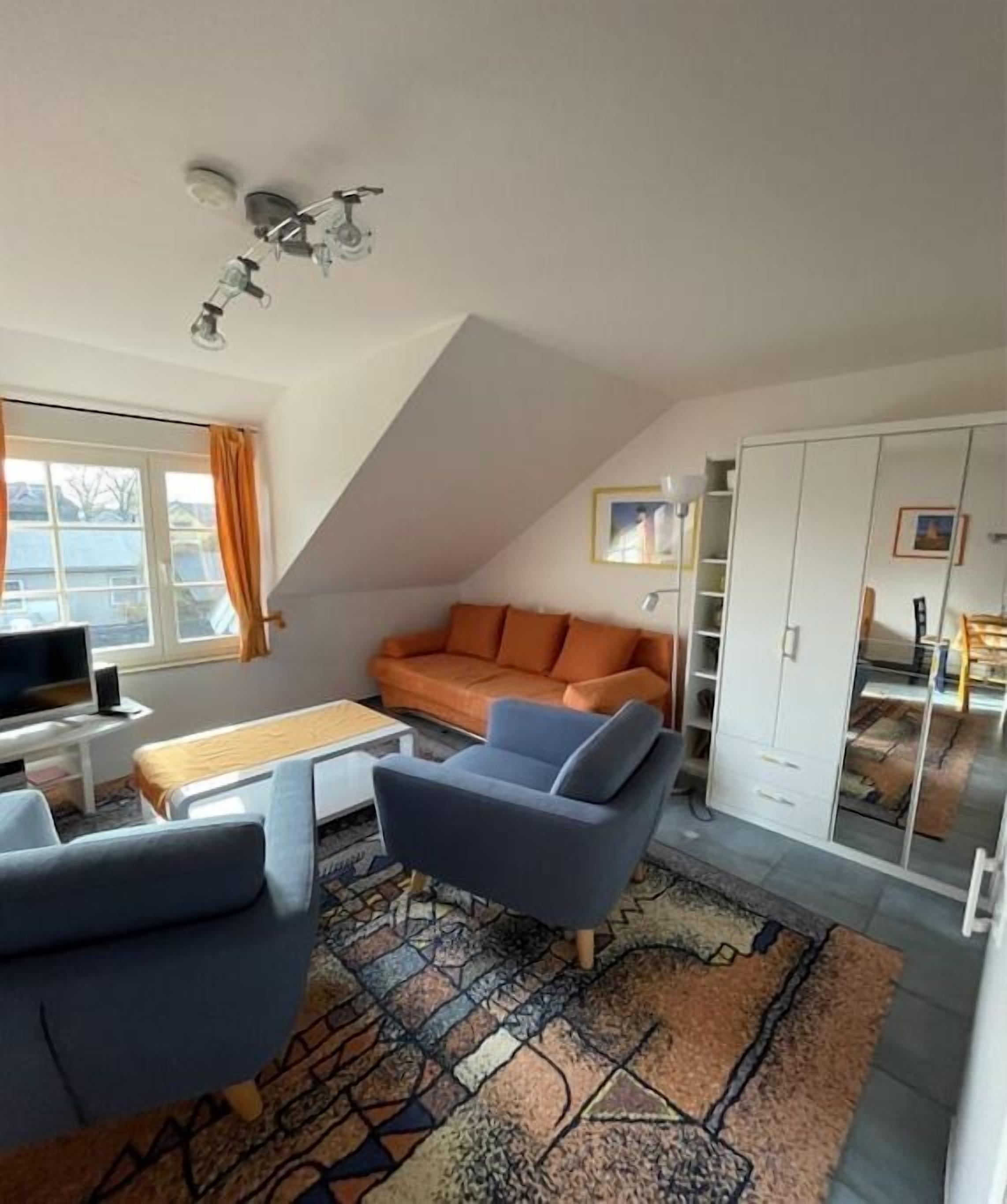 A modern living room with a sofa, a coffee table, and a television. There is a window with curtains, a patterned rug, and a wardrobe.