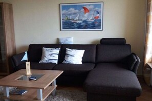 A living room with a dark-colored sofa, a coffee table with a remote control, and a painting of sailboats on the wall. - Holiday apartment in Damp with swimming pool (Damp)