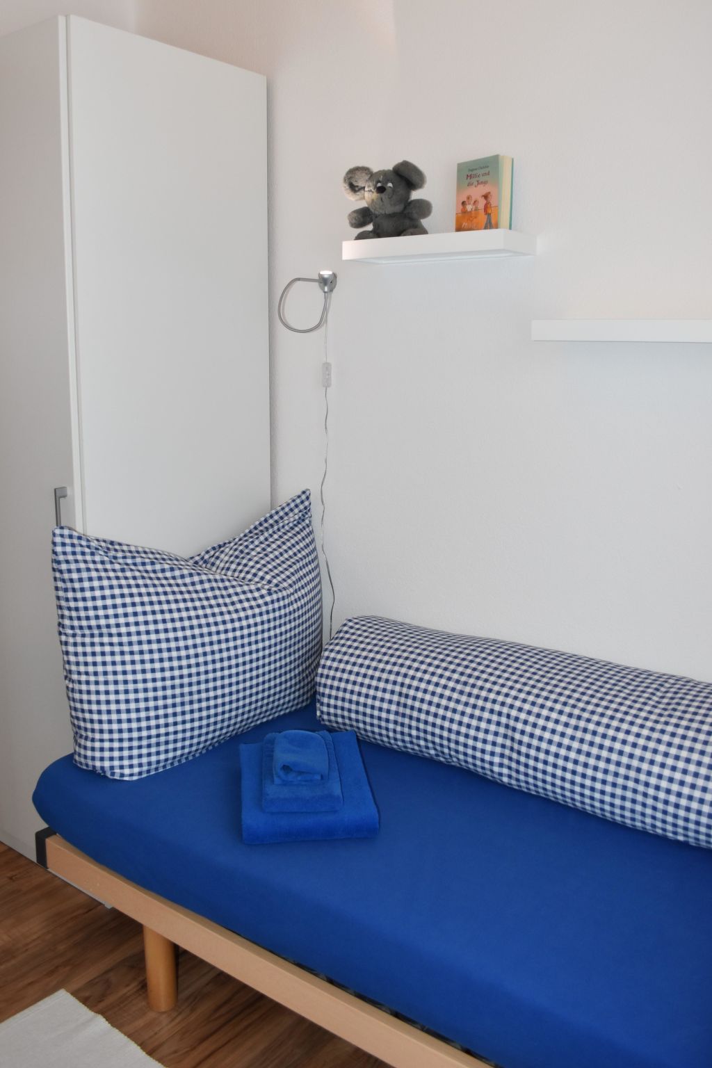 Ein Schlafzimmer mit einem Bett, Kissen, einem Nachttisch, einer an der Wand befestigten Ablage mit einem Buch und einem Teddybären sowie einer Tür.