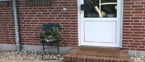 A brick house with a white door and windows, a small table with a plant, and a gravel driveway.
