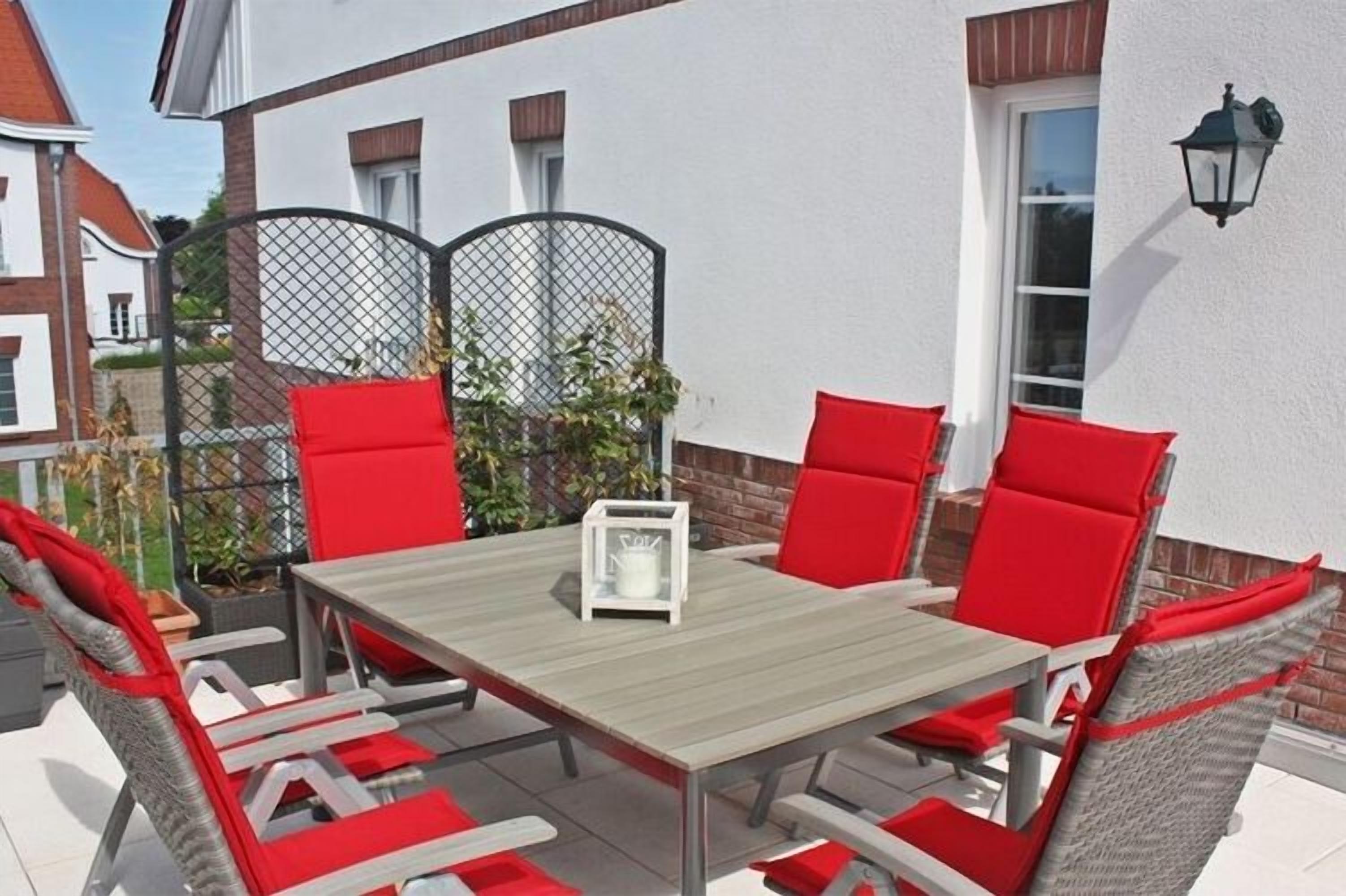 An outdoor patio with a wooden table and red cushioned chairs, a white coffee table, and a black lantern on the wall.