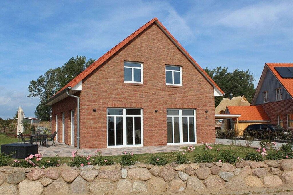Ein zweistöckiges Ziegelhaus mit geneigtem Dach, großen Fenstern und einer Steinmauer davor.