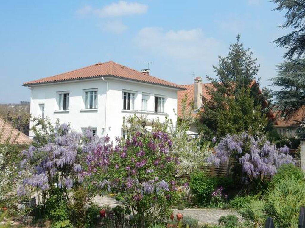 Une maison à deux étages, avec un toit recouvert de tuiles rouges et des murs blancs, entourée d’une végétation luxuriante et d’arbres aux fleurs violettes.