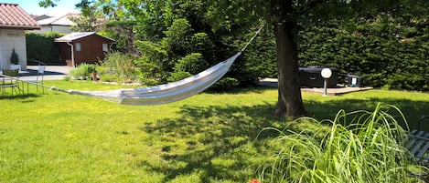 A hammock tied between two trees in a grassy area with a house and a shed in the background.