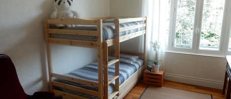 A bunk bed with a panda plush toy on top, a small bedside table with a plant, and a window with a view of trees.