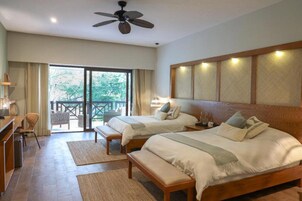 Habitación de hotel con dos camas, un escritorio, una silla, ventilador de techo y balcón con vistas a árboles.