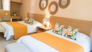 A hotel room with two beds, each with a white and orange bedspread, and matching floral pillows. There is a wooden nightstand with a lamp and a small table with a glass.