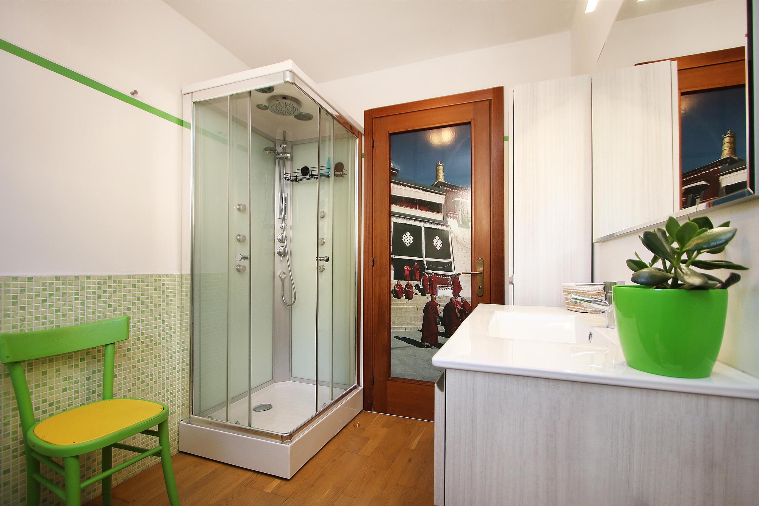 Ein modernes Badezimmer mit einer gläsernen Duschkabine, einem grünen Stuhl, einem Waschbecken mit einer grünen Pflanze und Blick auf ein Gebäude mit Menschen draußen.