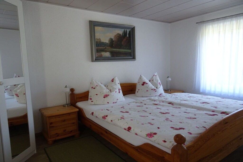 Un dormitorio con una cama de madera, ropa de cama blanca, una mesita de noche, una lámpara, un cuadro en la pared y una ventana con cortinas.