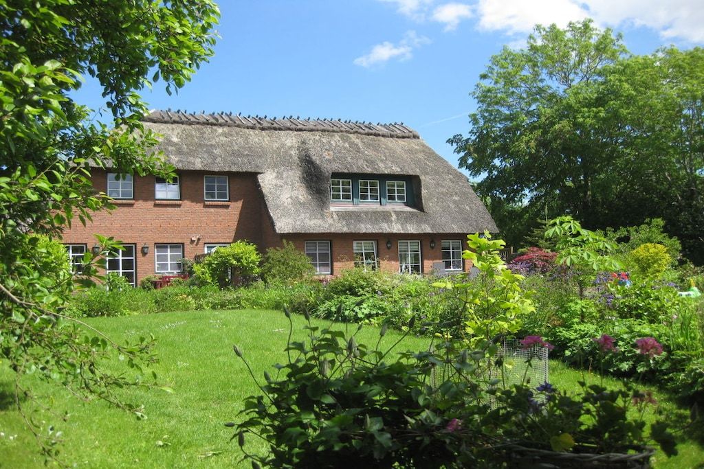 A traditional brick house with a thatched roof, surrounded by a well-maintained garden with various plants and flowers.