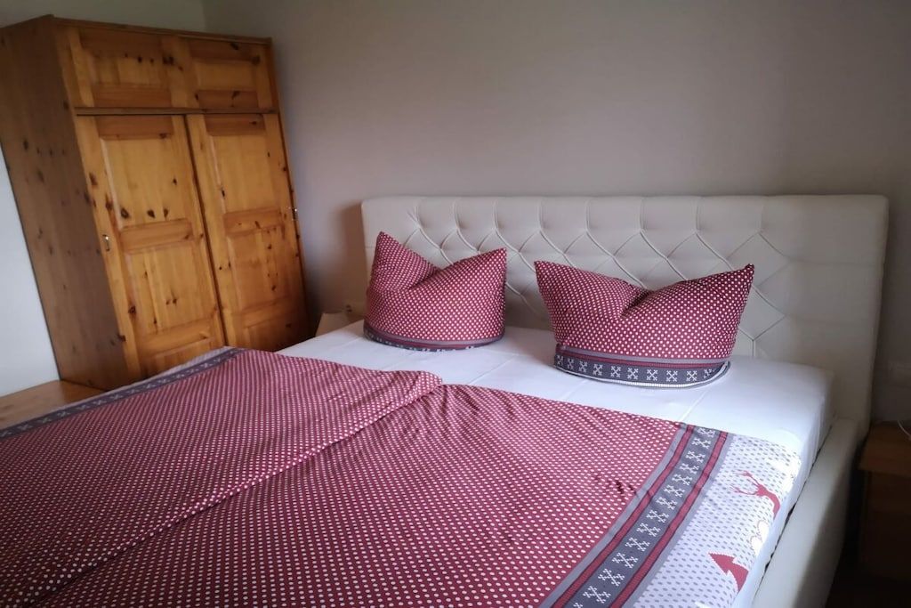 Une chambre à coucher avec une tête de lit blanche recouverte de tissu, deux coussins rouges, une armoire en bois et une couvre-lit à motifs.