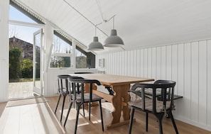 Et spiseområde med et træbord og sorte stole, hvide panelvægge og udsigt gennem en glasdør.
