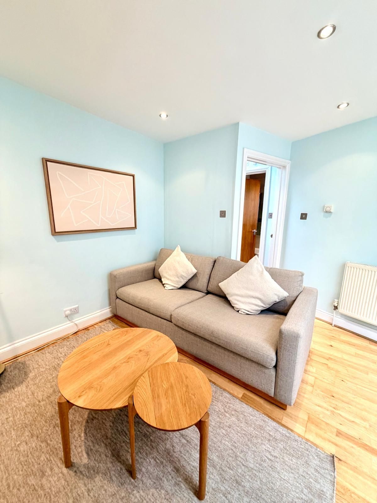 A living room with a grey sofa, two wooden side tables, a framed abstract artwork, and a radiator.