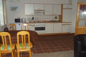 A compact kitchen with white cabinets, a microwave, a stove, and a sink. There are two wooden chairs with cushions and a dining table. - Island apartment Fehmarn (Puttgarden\/Fehmarn)