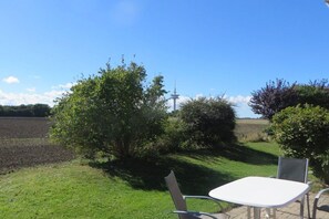 An outdoor seating area with a table and chairs, surrounded by greenery and a clear sky. - Island apartment Fehmarn (Puttgarden\/Fehmarn)