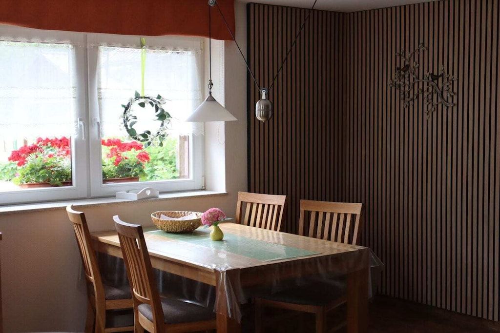A dining area with a table, chairs, a hanging light fixture, and a window with a view of flowers.