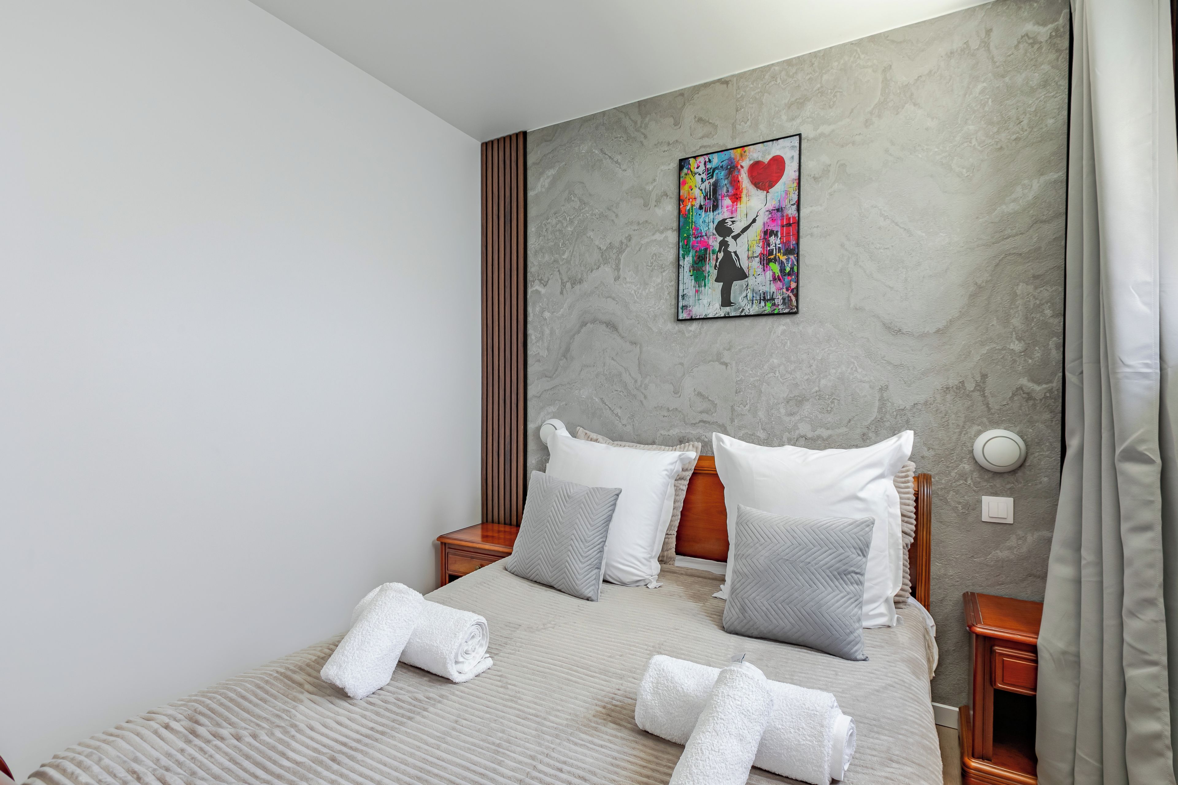 A hotel room with a bed, pillows, a headboard, a nightstand, a wall-mounted painting, and a curtain.