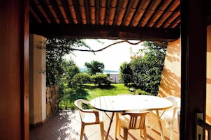 Una terrazza esterna con un tavolo rotondo e sedie, un pergolato in legno sopra e vista su un giardino con alberi e uno specchio d'acqua in lontananza.