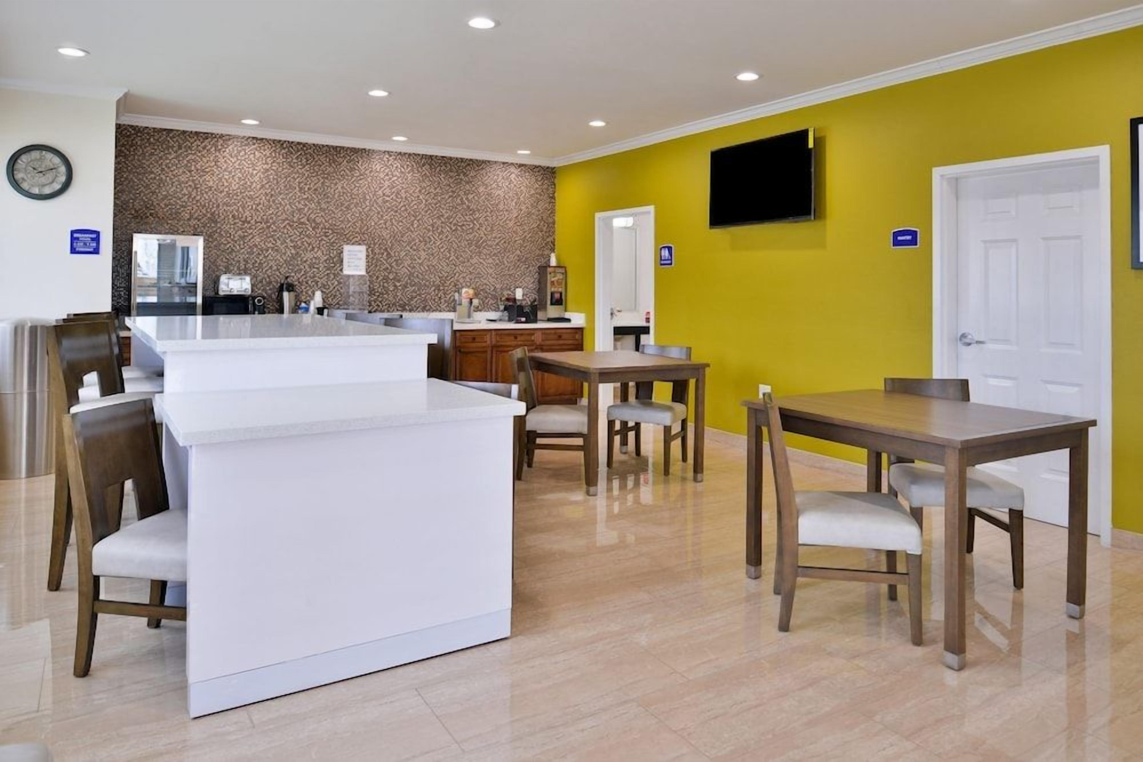 A hotel room with a kitchenette, a dining area with a table and chairs, a TV mounted on the wall, and a door with a sign.