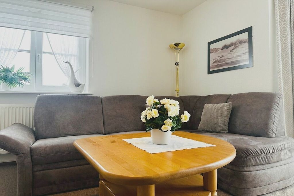 A living room with a grey sofa, a wooden coffee table with a white cloth, a potted plant, a floor lamp, and a framed picture on the wall.