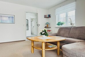 A living room with a round wooden coffee table, a grey sofa, a bookshelf, a window with a plant, and a door leading to another room. - Holiday home with 1 bedroom (St. Peter - Ording)