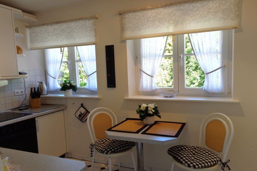 Una cocina con dos ventanas, una mesa de comedor con sillas y una pequeña encimera con un hervidor.
