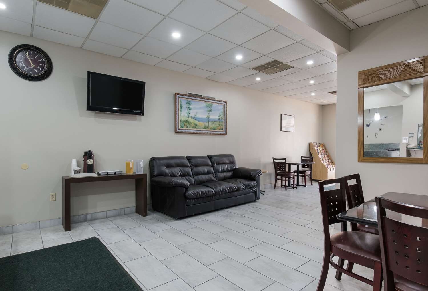 A living room with a black leather sofa, a coffee station, a flat-screen TV, a framed landscape painting, and a dining area with chairs and a table.