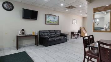 A living room with a black leather sofa, a coffee station, a flat-screen TV, a framed landscape painting, and a dining area with chairs and a table.