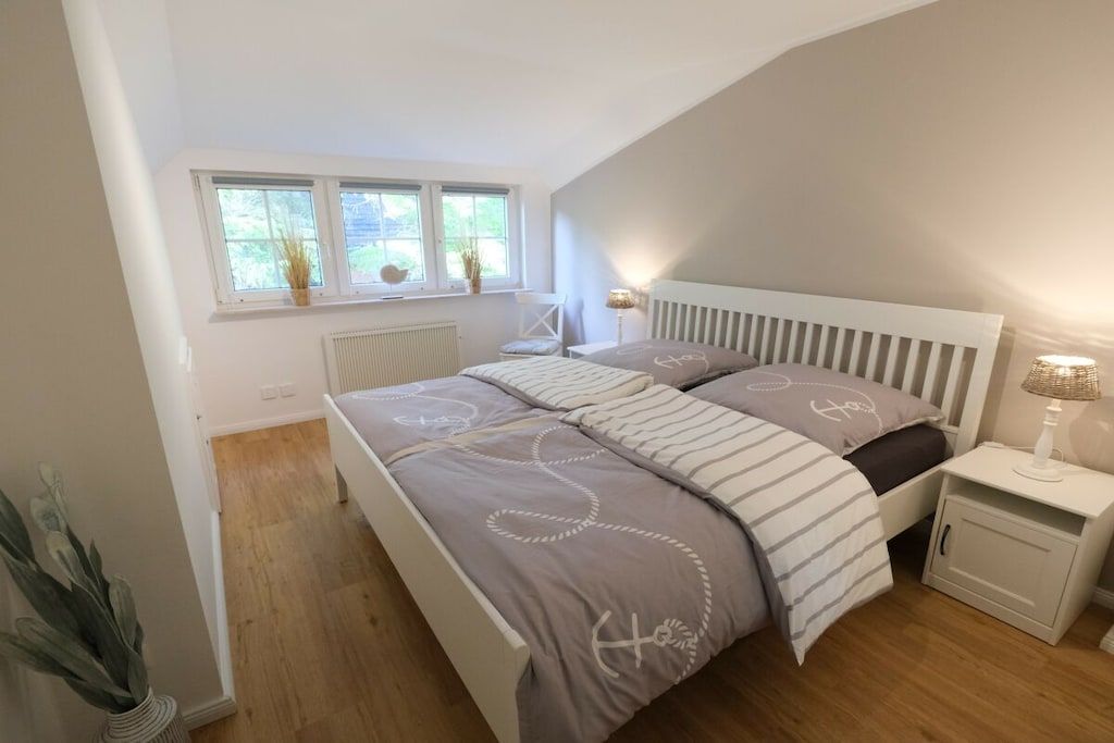 Ein Schlafzimmer mit Bett, Nachttisch, Lampe, Heizkörper, Fenster und einer Pflanze.