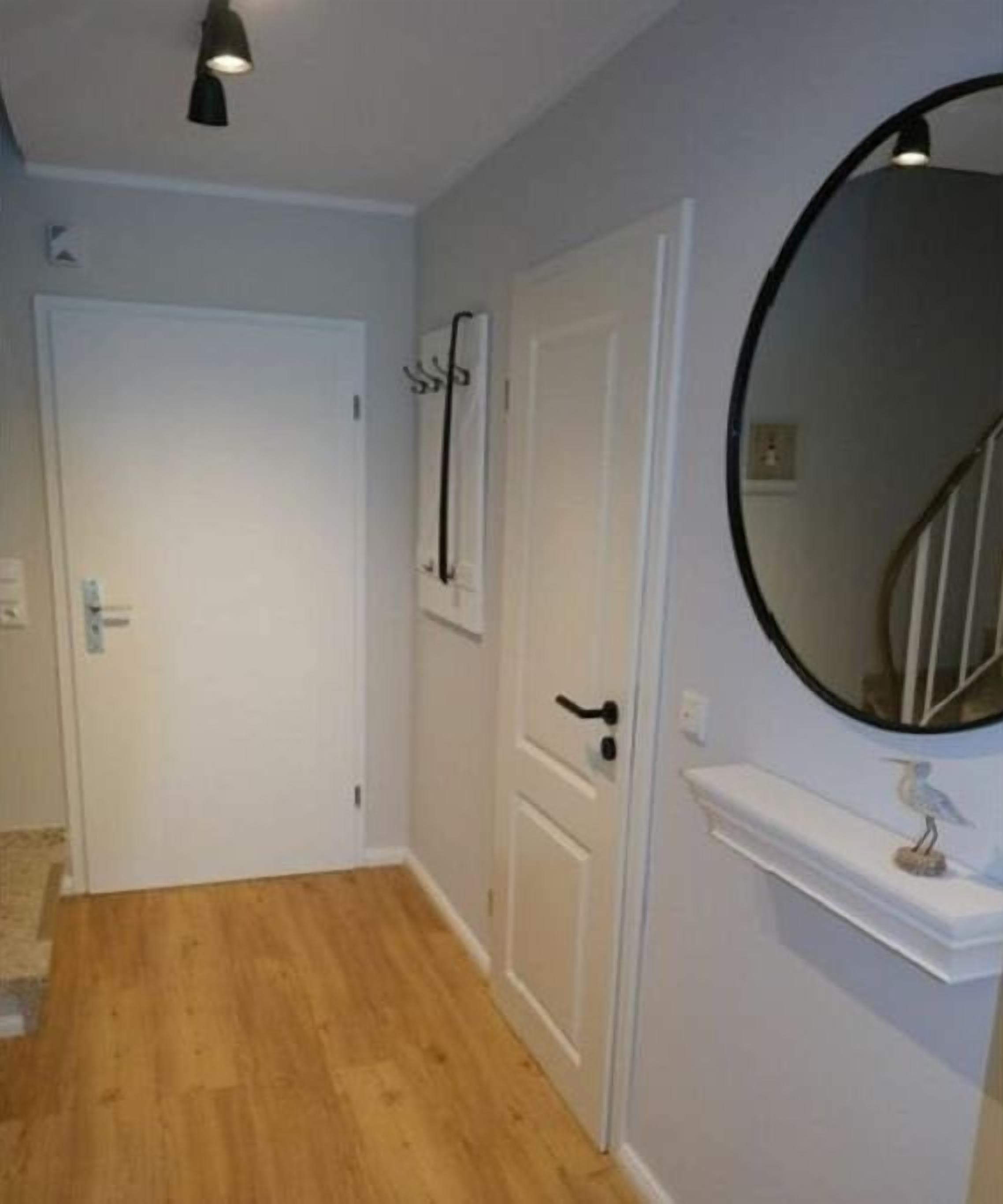 Een gang met houten vloer, witte deuren, een wit wandplankje met een klein sculptuur en een ronde spiegel.