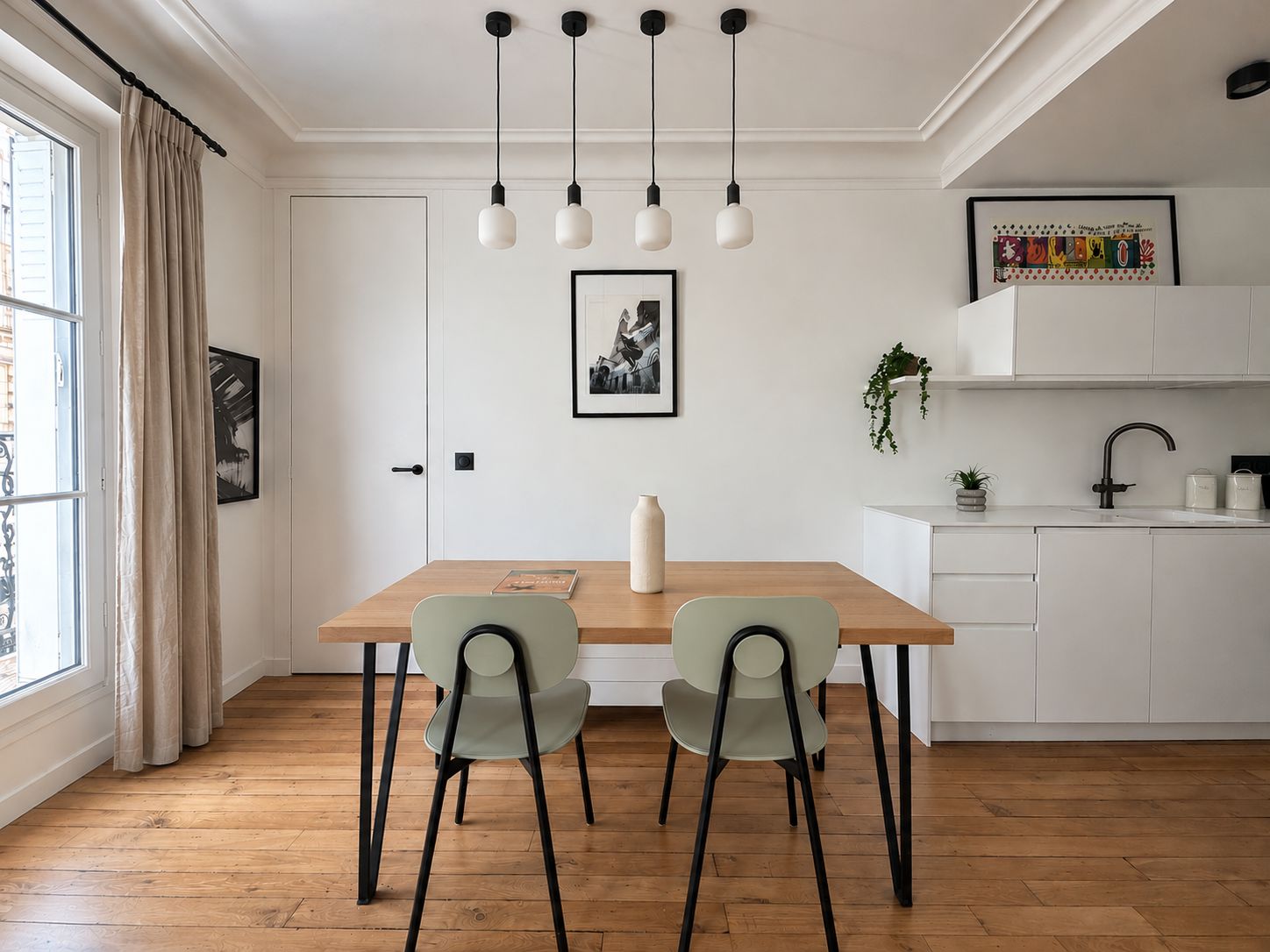 A dining area with a wooden table, two chairs, a hanging light fixture, a framed picture on the wall, and a kitchen area with a sink and cabinets.